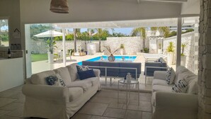 A spacious living area with a white sofa, a coffee table, and a view of a swimming pool and garden.