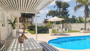 A covered outdoor area with a pool, white furniture, and potted plants.