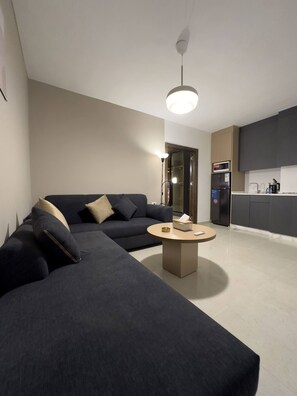 A modern living room with a dark sofa, a round coffee table, and a kitchen area with cabinets and appliances.