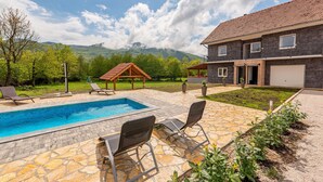 Una piscina con lettini, una zona relax coperta e una casa con facciata in pietra e tetto a tegole.