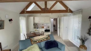 A spacious living area with a blue sofa, wooden ceiling beams, a dining table, and a kitchen in the background.