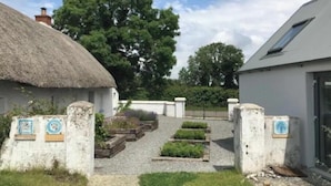 A rural property with a thatched-roof house, a modern building, and a gravel pathway leading to a gate with plaques.