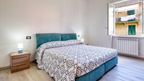 Um quarto com uma cama, duas mesas de cabeceira, uma janela com vista para prédios e um radiador.