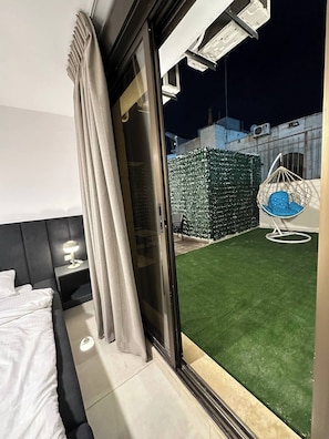 A hotel room with a bed, a nightstand, a lamp, a sliding glass door to an outdoor area with artificial grass and a hanging chair. - Stylish Luxe Apartment (Amman)