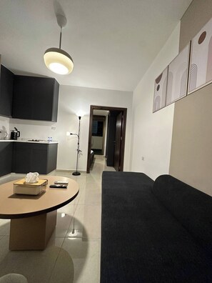 A modern living room with a dark sofa, a round coffee table, and a kitchen area with cabinets and a stove. - Stylish Luxe Apartment (Amman)