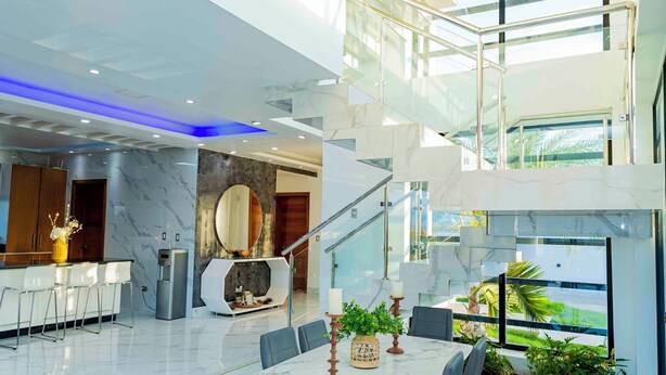 Un interior moderno con piso de baldosas de mármol, una mesa de comedor central con sillas, una escalera con barandas de cristal y vistas al exterior con vegetación.