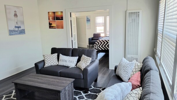 A modern living room with a black leather sofa, a wooden coffee table, and a patterned rug. There is a door leading to another room with a bed and a chair. - Modern 2 bed 2 bath Paradise - Mins. from LAX & SOFI (Los Angeles)