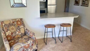 Une petite cuisine avec un plan de travail blanc, deux tabourets et un fauteuil à motifs.