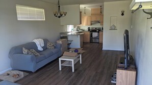 A living room with a sofa, a coffee table, and a television. There is a kitchen in the background with cabinets and a stove.