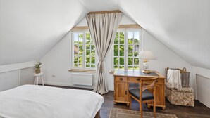 Une chambre avec un plafond en pente, un lit, un bureau avec une chaise, une lampe, un radiateur et une fenêtre avec des rideaux.