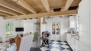 Une pièce dotée d’un sol à carreaux, de poutres en bois au plafond, d’une table à manger avec des chaises, d’un coin cuisine avec des meubles blancs et d’une grande fenêtre à carreaux multiples.