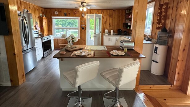 A wooden cabin kitchen with a central island, bar stools, a refrigerator, a stove, and a window with a view of trees. - Lake Rosseau Water's Edge Retreat (Port Carling)