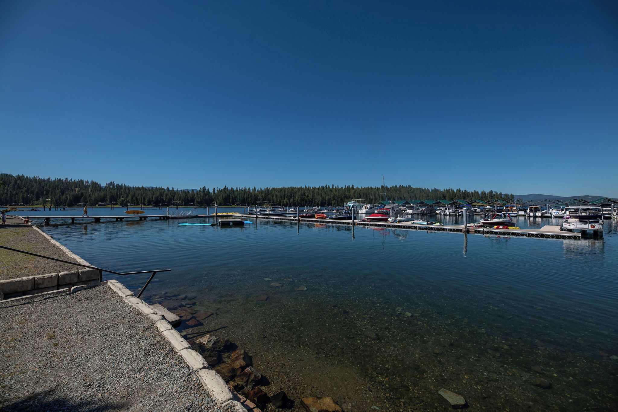 A marina with boats docked, clear water, and a forested hillside in the background.