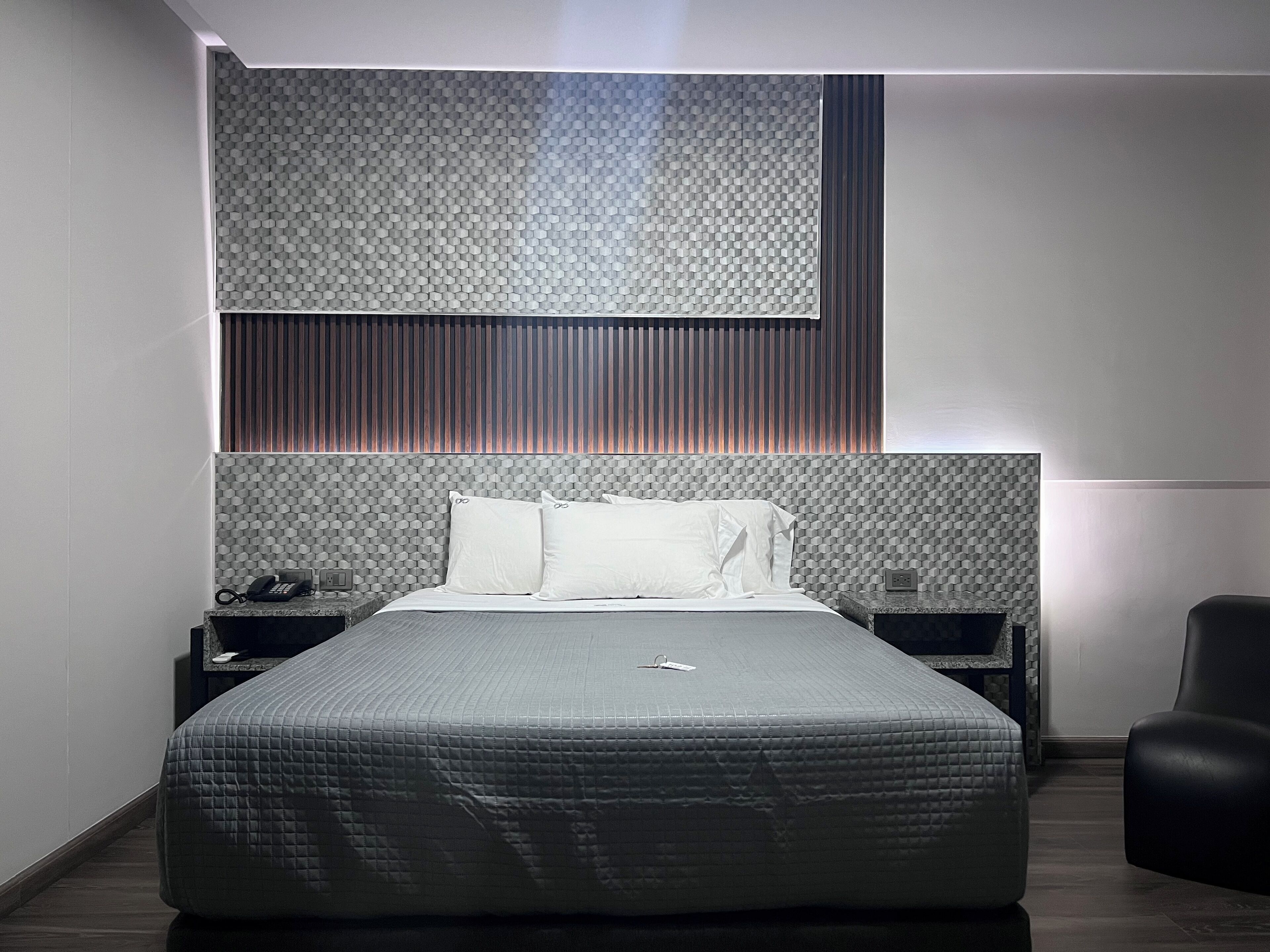 A modern bedroom with a large bed, a headboard with a textured pattern, a bedside table with a phone, and a chair.