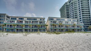 A beachfront hotel with multiple balconies, a sandy beach in the foreground, and a tall building in the background. - Panama City Beach Condo w/ King Suite & Pool View (Panama City Beach)