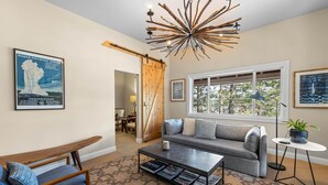 A living room with a sofa, coffee table, and a unique chandelier. There is a large window and a door leading to another room. - Monthly Rental - Crimson 4 BR Cabin By Tahoe Getaways - Walk Downtown Truckee (Truckee)