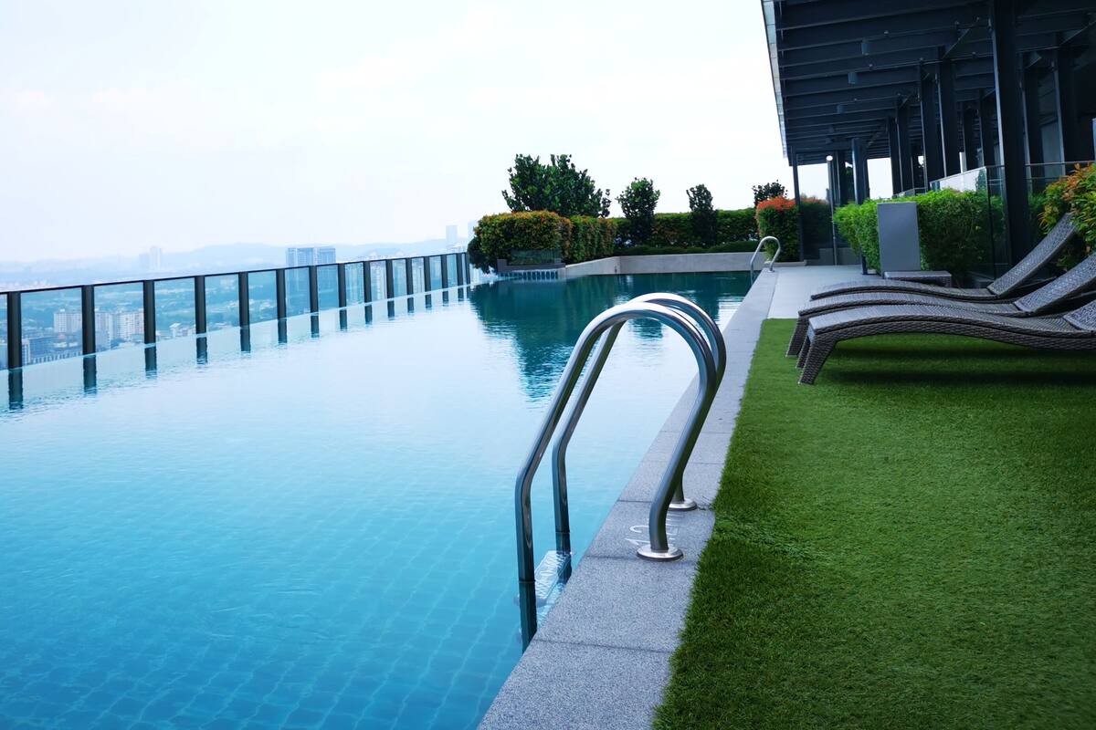 A rooftop infinity pool with a glass barrier, a grassy area with lounge chairs, and a cityscape view in the background.