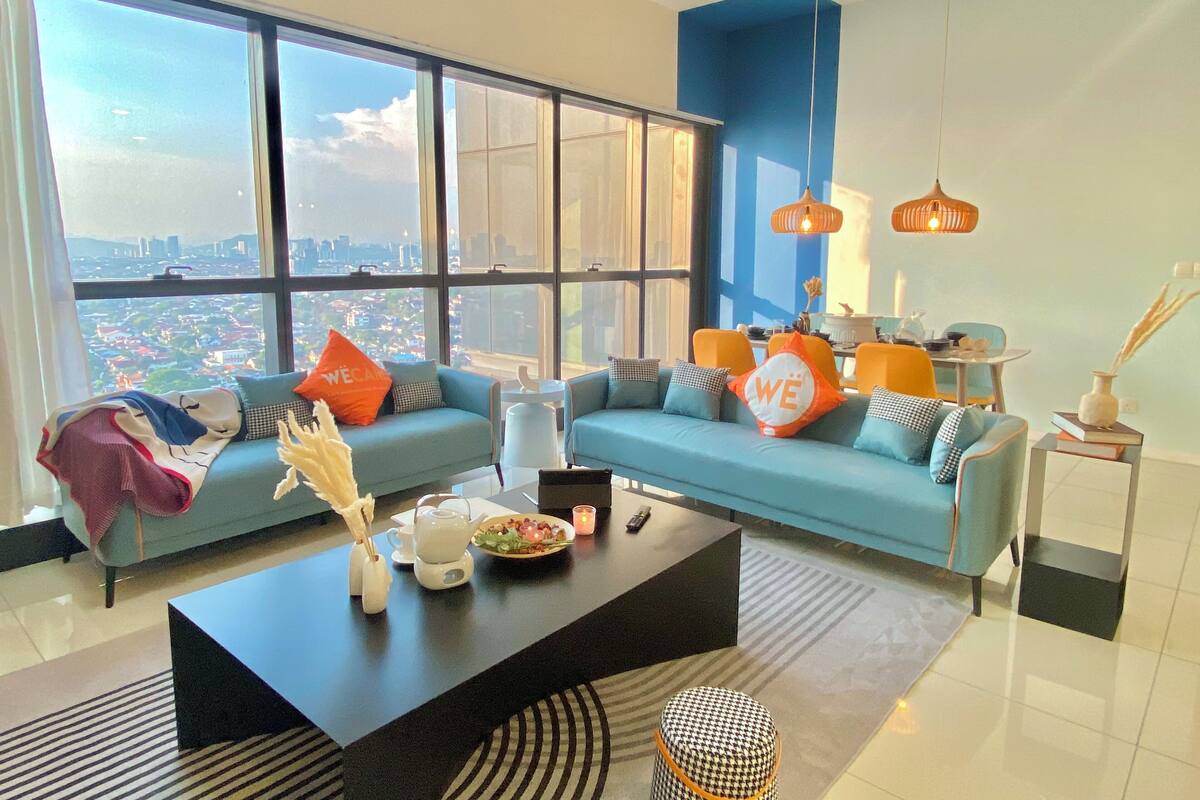 A modern living room with a large window, a blue sofa set, a coffee table, and a dining area with orange chairs.