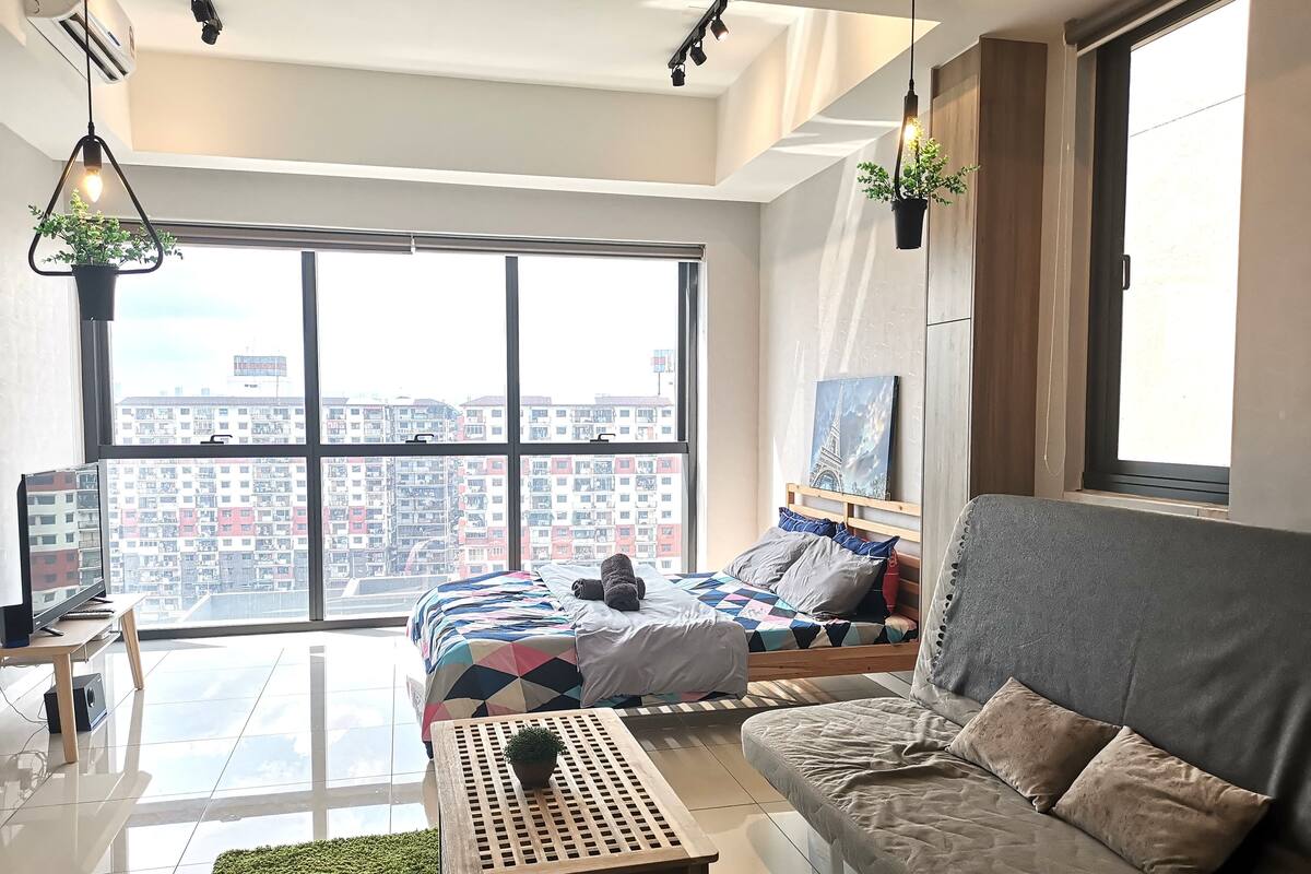A modern apartment with a large window, a bed with a colorful quilt, a grey sofa, and a small table with a plant.