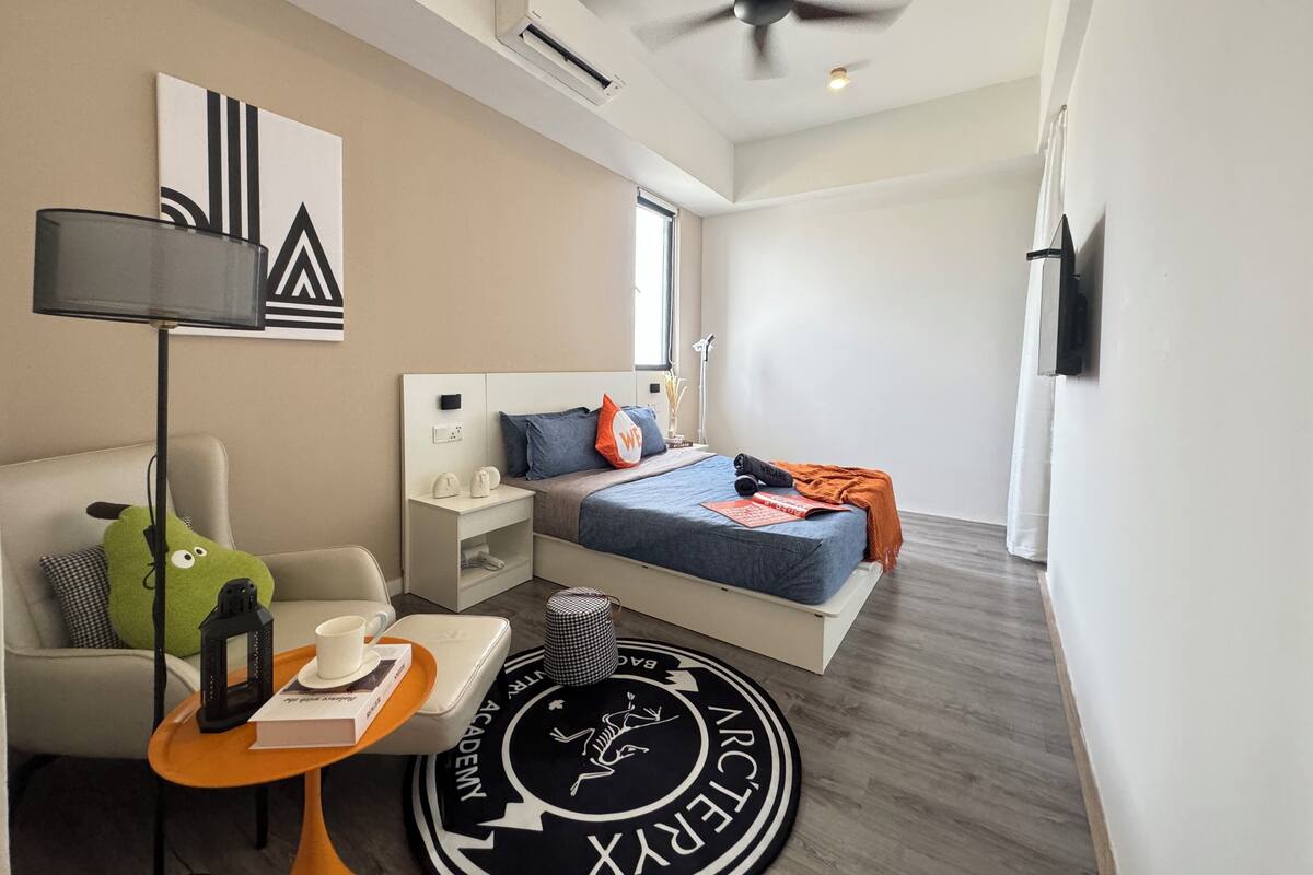 A modern hotel room with a bed, bedside table, lamp, and a round rug with a logo.