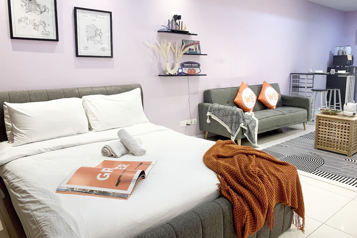A modern bedroom with a large bed, grey sofa, and a small kitchen area.