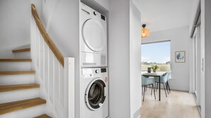 A modern laundry room with stacked washers and dryers, a wooden staircase, and a view of the outdoors through a large window.
