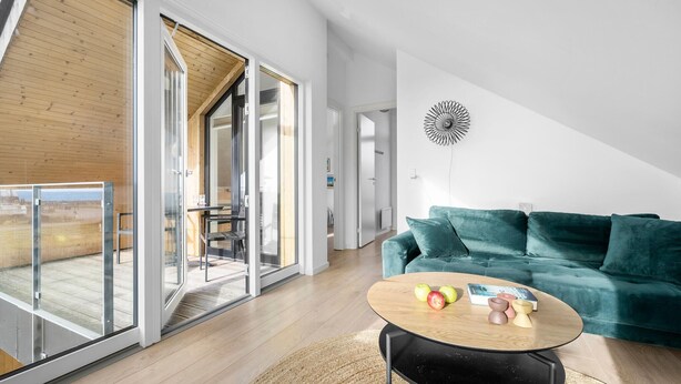 A modern living room with a teal sofa, a round coffee table with apples and a book, and a glass door leading to an outdoor area.