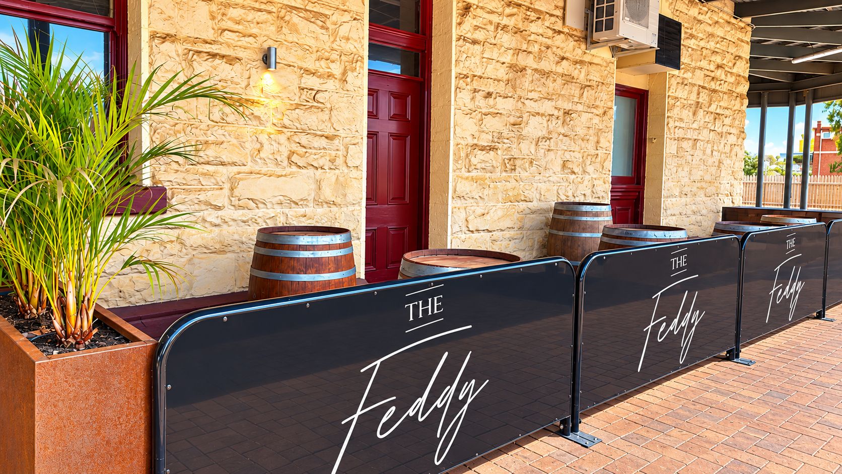 Un bâtiment en briques avec une porte rouge, des tables à l’extérieur et des barils servant de sièges, ainsi qu’une enseigne sur laquelle on peut lire « THE Feddy ».