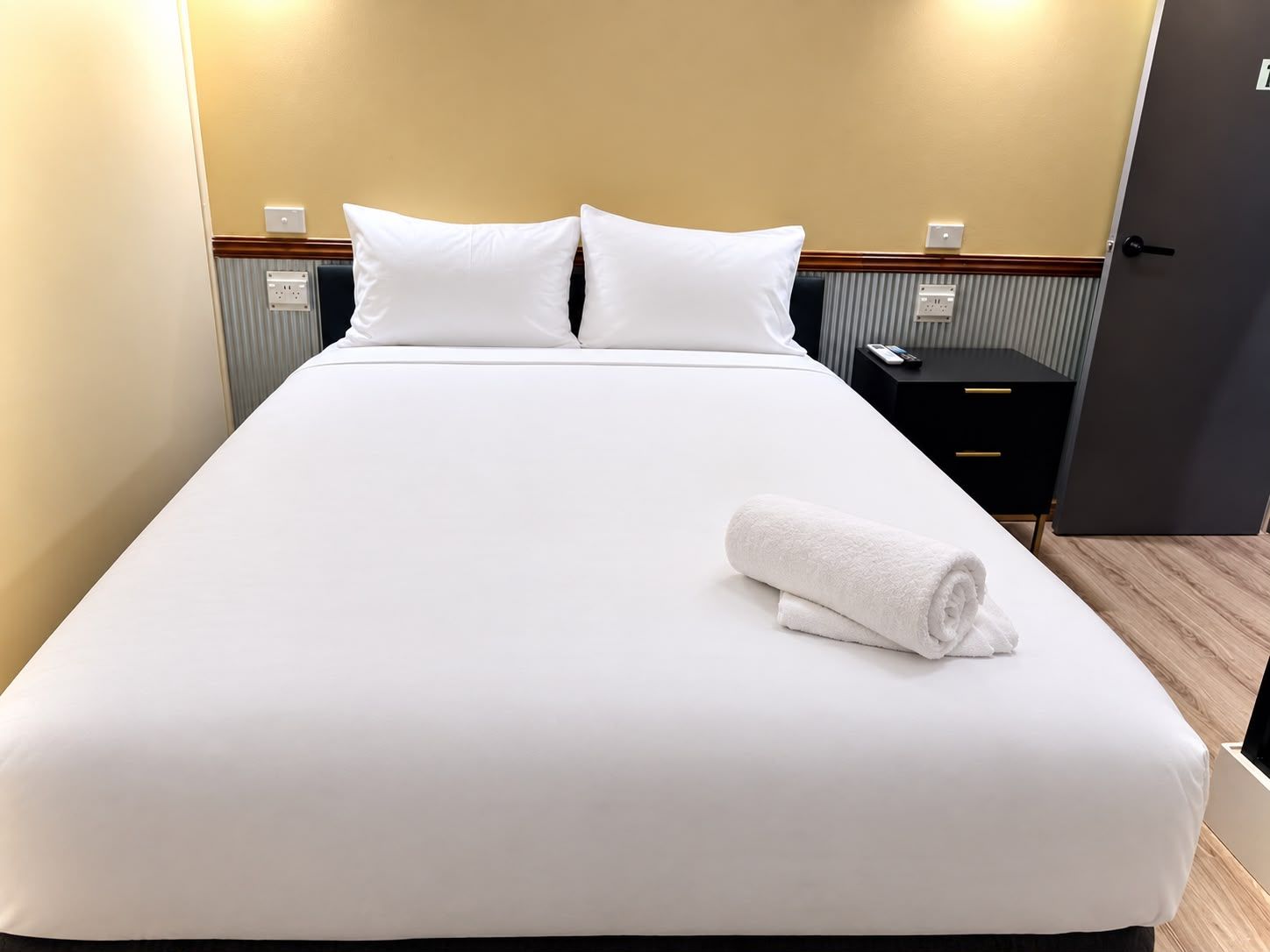 A neatly made bed with white linens and pillows, a rolled white towel, and a black nightstand with a remote control.