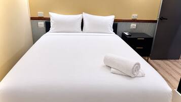 A neatly made bed with white linens and pillows, a rolled white towel, and a black nightstand with a remote control.