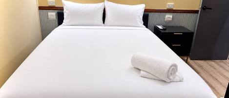 A neatly made bed with white linens and pillows, a rolled white towel, and a black nightstand with a remote control.