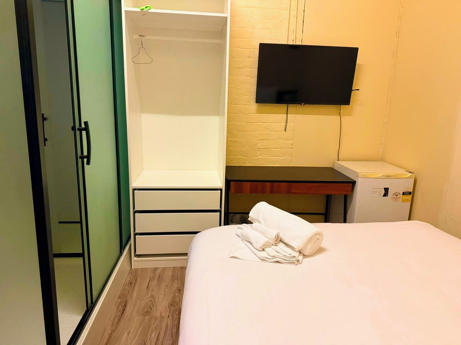 A hotel room with a bed, a wardrobe, a desk with a flat-screen TV, and a small refrigerator.