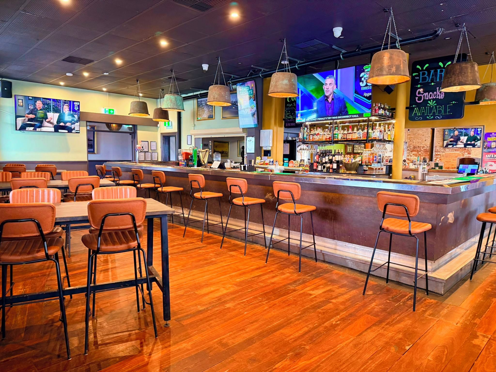 A bar area with a long counter, multiple bar stools, and a television screen displaying a sports event.