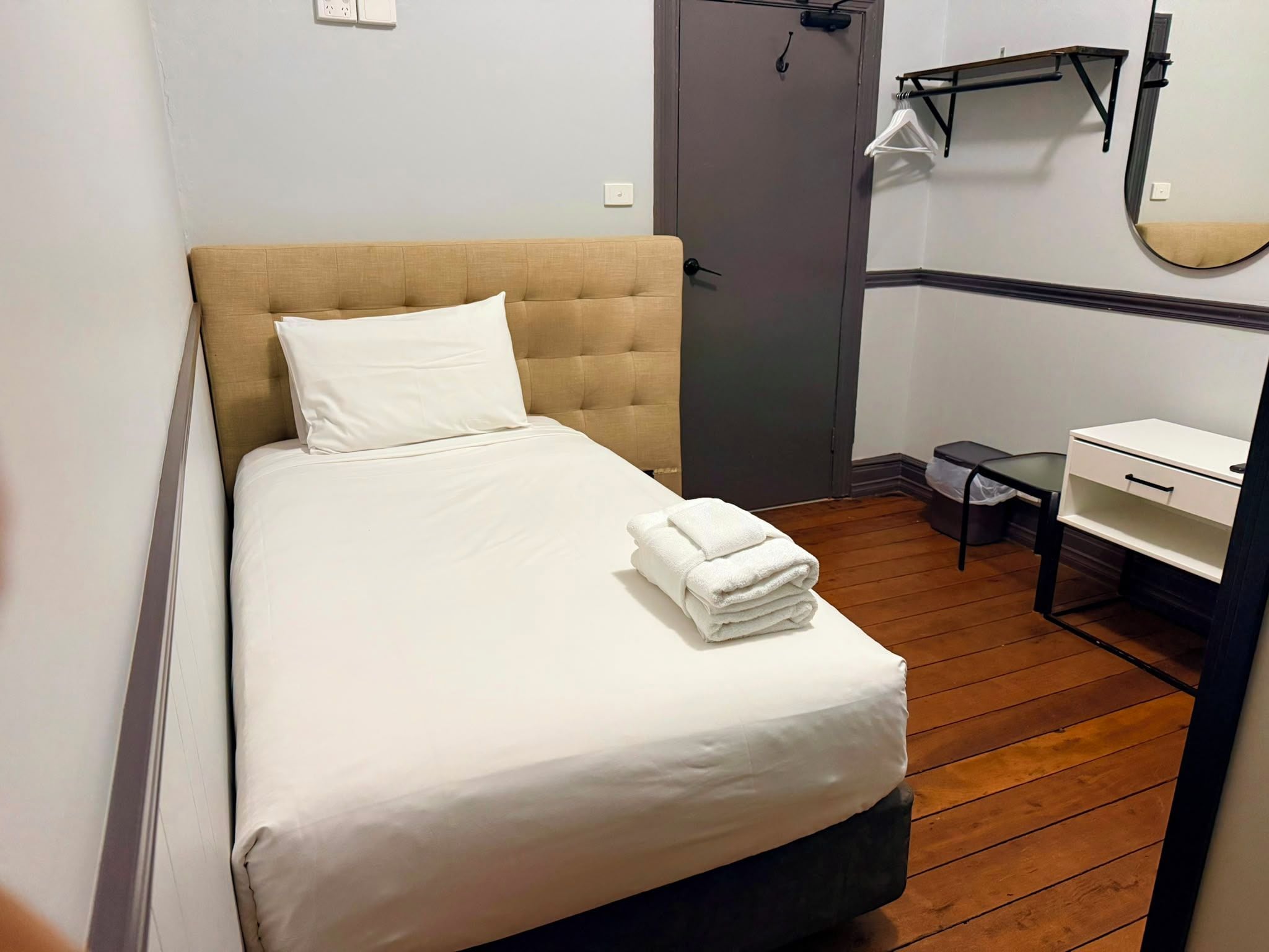 A single bed with a headboard, a bedside table, a small desk with a chair, a mirror, and a door.