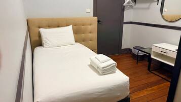 A single bed with a headboard, a bedside table, a small desk with a chair, a mirror, and a door.