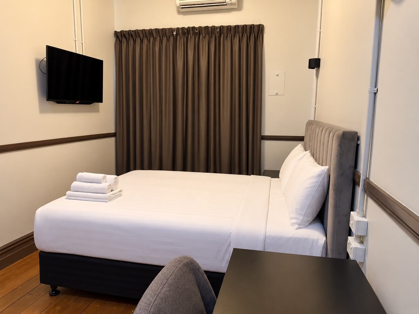 A hotel room with a bed, a flat-screen TV mounted on the wall, a window with curtains, and a bedside table.