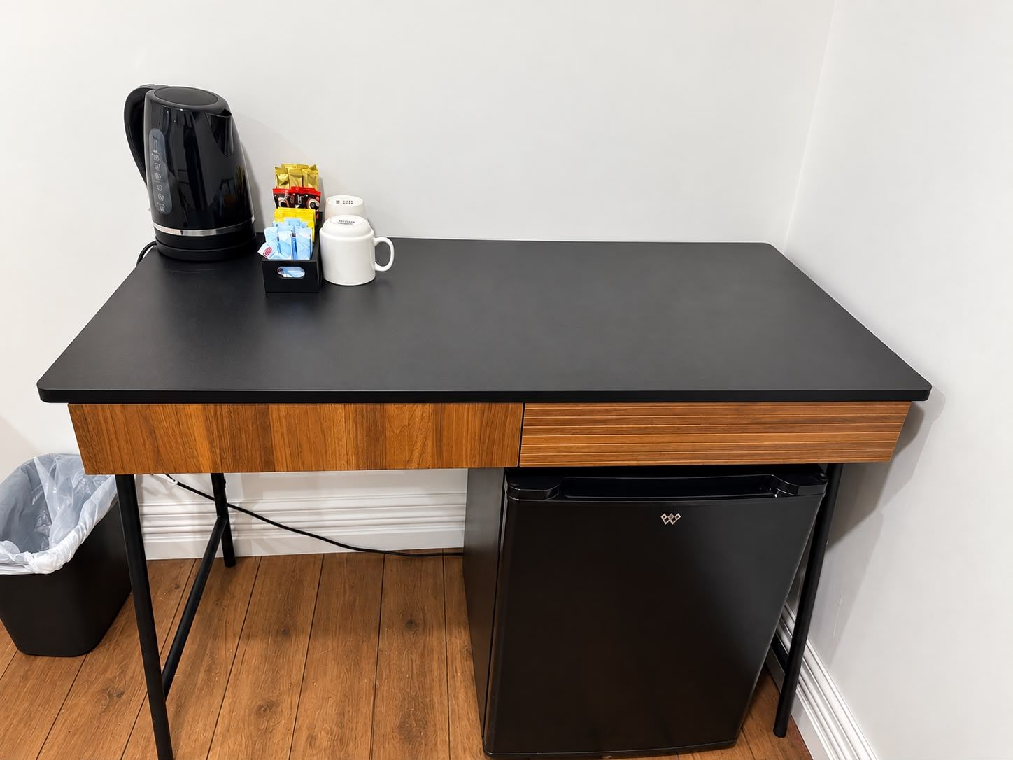A compact kitchenette with a black countertop, a coffee maker, a mug, and a trash bin.