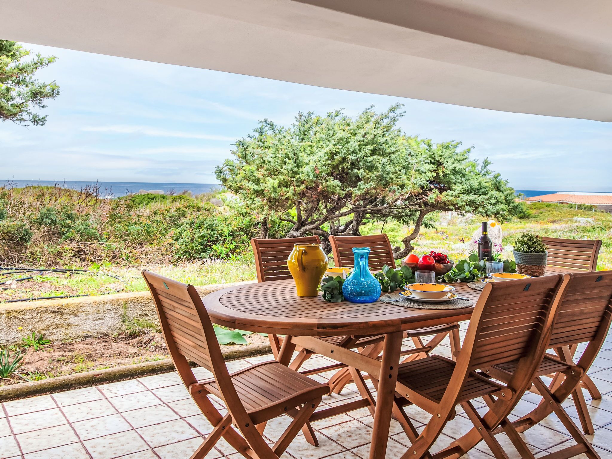A round wooden table with chairs, set on a tiled patio overlooking a scenic landscape with trees and a view of the sea.