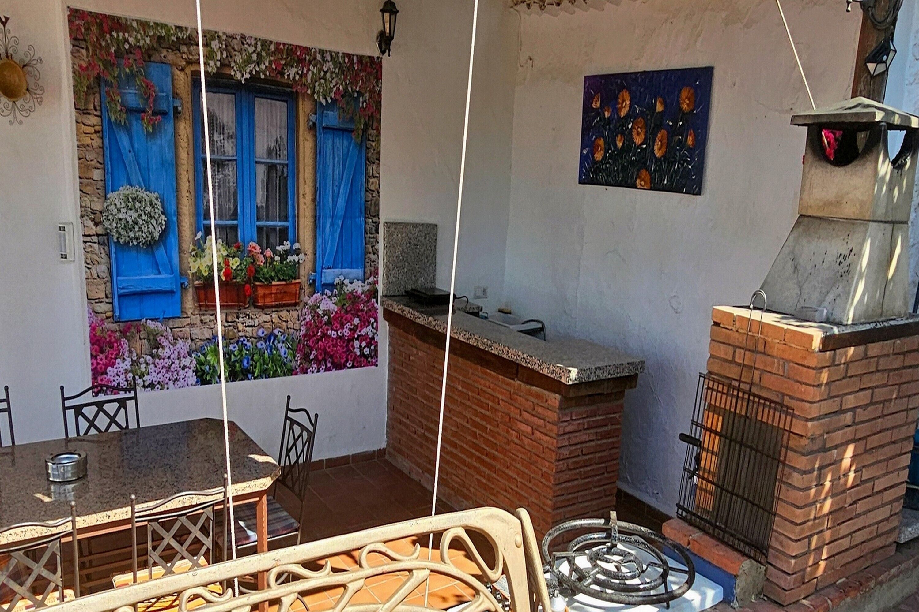 Een terras met een tafel en stoelen, een blauw raam met gordijnen, een bloemenwand en een bakoven.