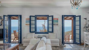 A room with a white sofa, a wooden coffee table, and a view through open doors to the sea. - Country house "Agios Sostis" with a jacuzzi and close to the beach (Serifos)