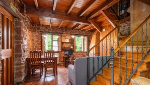 Un interior rústico con pared de piedra, escalera de madera y zona de comedor con sillas y una mesa.