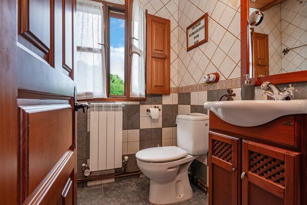 Un baño con inodoro, lavamanos, armarios de madera, una ventana con cortinas y una pared con azulejos.