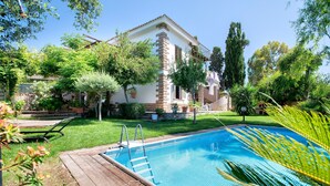 Une piscine dotée d’une terrasse en bois, entourée d’une végétation luxuriante et bordée d’un bâtiment au toit recouvert de tuiles.