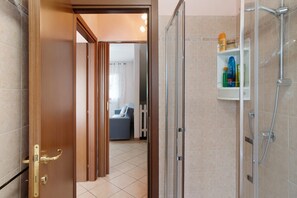 A bathroom with a glass shower enclosure, a white shelf with toiletries, and a wooden door leading to a living area with a sofa and curtains.