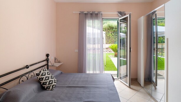 A bedroom with a bed, a pillow, a curtain, a sliding glass door leading to a balcony, and a small table. - Villa 'Il Frutteto 49' with Private Terrace and Wi-Fi (Ortovero)