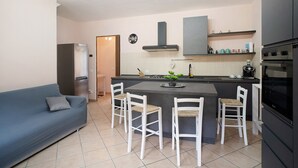 A compact kitchen with a central island, white chairs, a grey sofa, and a built-in oven. - Villa 'Il Frutteto 49' with Private Terrace and Wi-Fi (Ortovero)