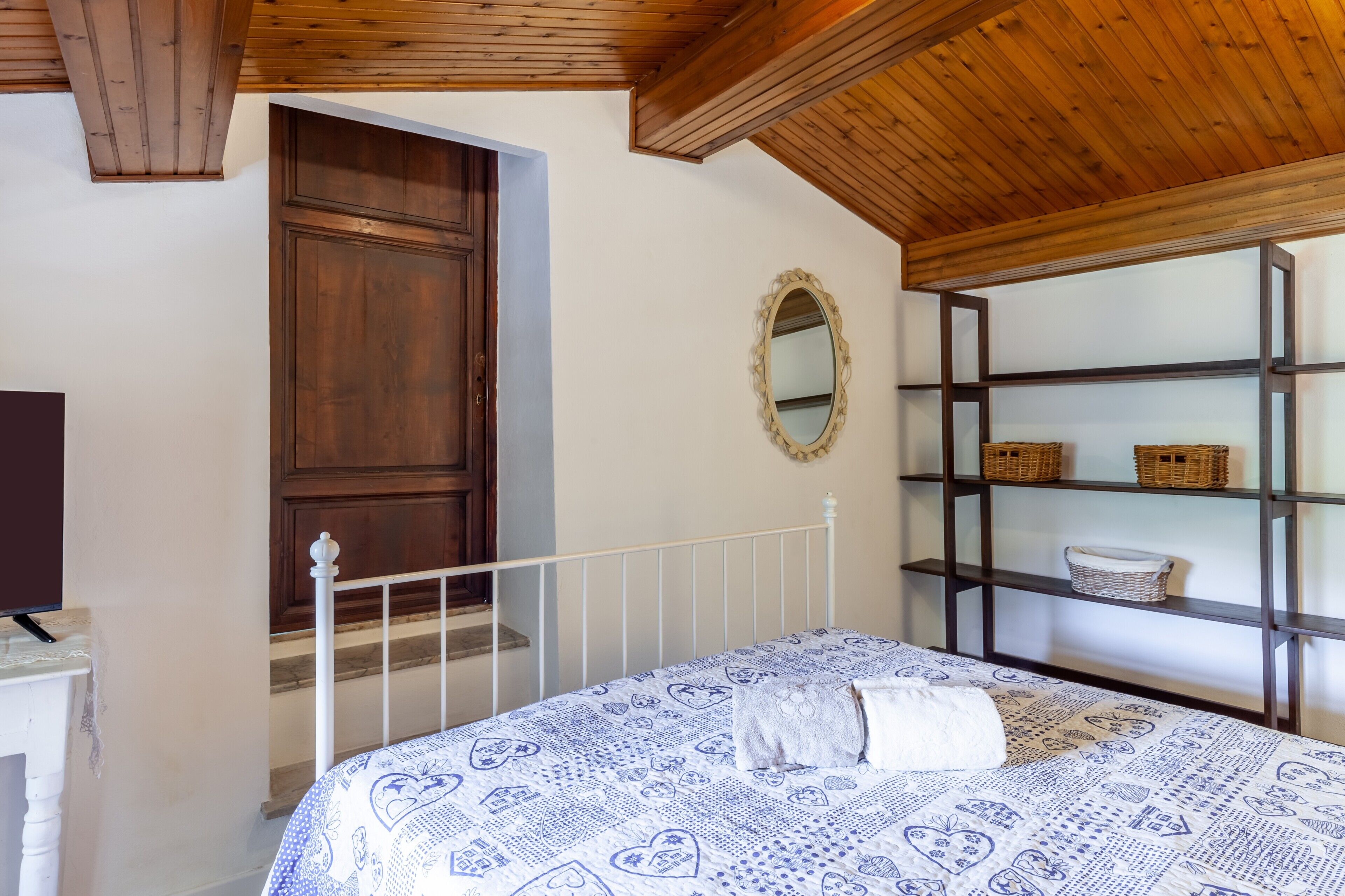 Une chambre avec un plafond en bois, un lit blanc avec une literie à motifs, une armoire en bois, un miroir et des étagères avec des paniers.