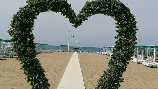 Uma praia com um arco em forma de coração feito de vegetação, um caminho branco e sacos de feijão listrados em vermelho e branco.