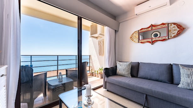 Una sala de estar moderna con balcón que da al mar, un sofá, una mesa de centro de vidrio y un espejo decorativo.