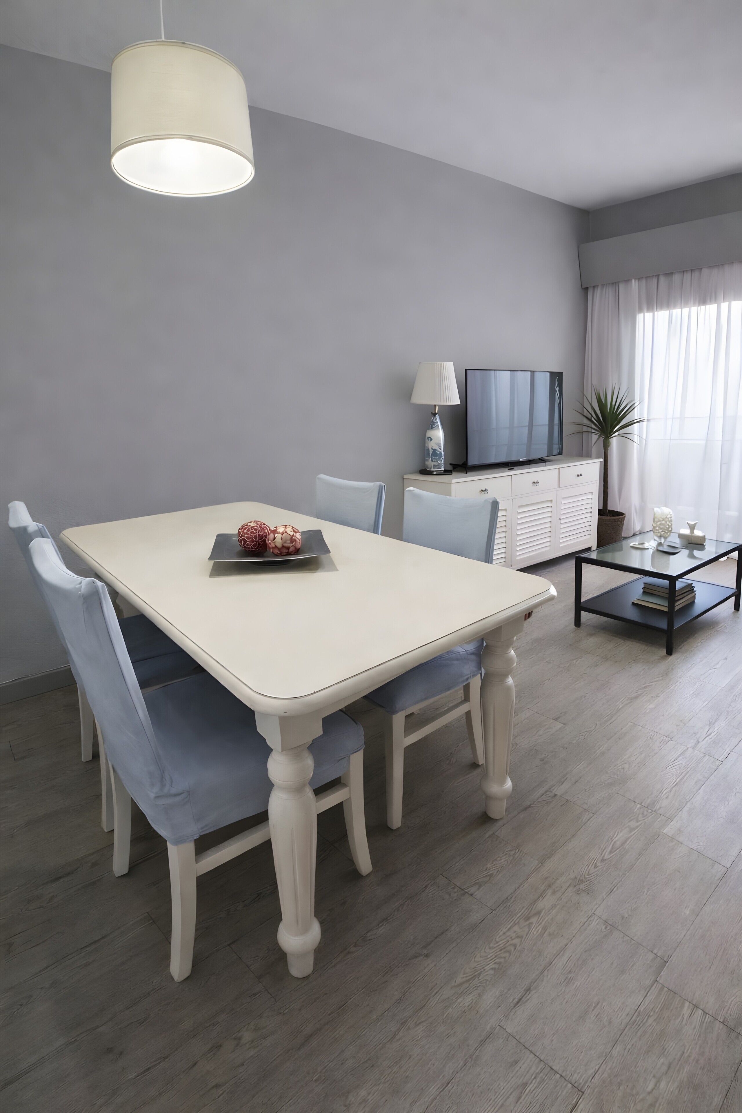 A dining area with a white table, blue chairs, a hanging light fixture, a television on a cabinet, and a potted plant.